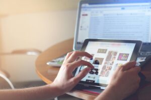 Hands using a tablet beside a laptop in a home office setting, showcasing online browsing.